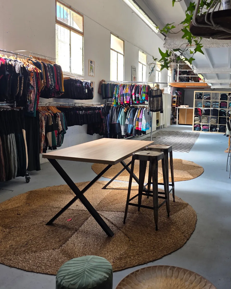Inside the La Gata Maula showroom: clothing rails, a wooden table with metal stools on natural-fibre rugs, and shelves of folded garments.